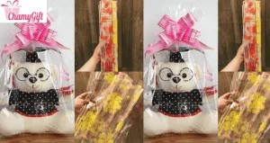 Một chú gấu bông màu kem được gói cẩn thận trong hộp quà carton màu hồng nhạt, thắt nơ ruy băng trắng, đặt trên nền gỗ mộc mạc.